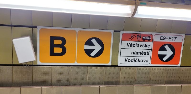 Yellow Prague Metro Line B sign with a right-pointing arrow beside signs for street exits.